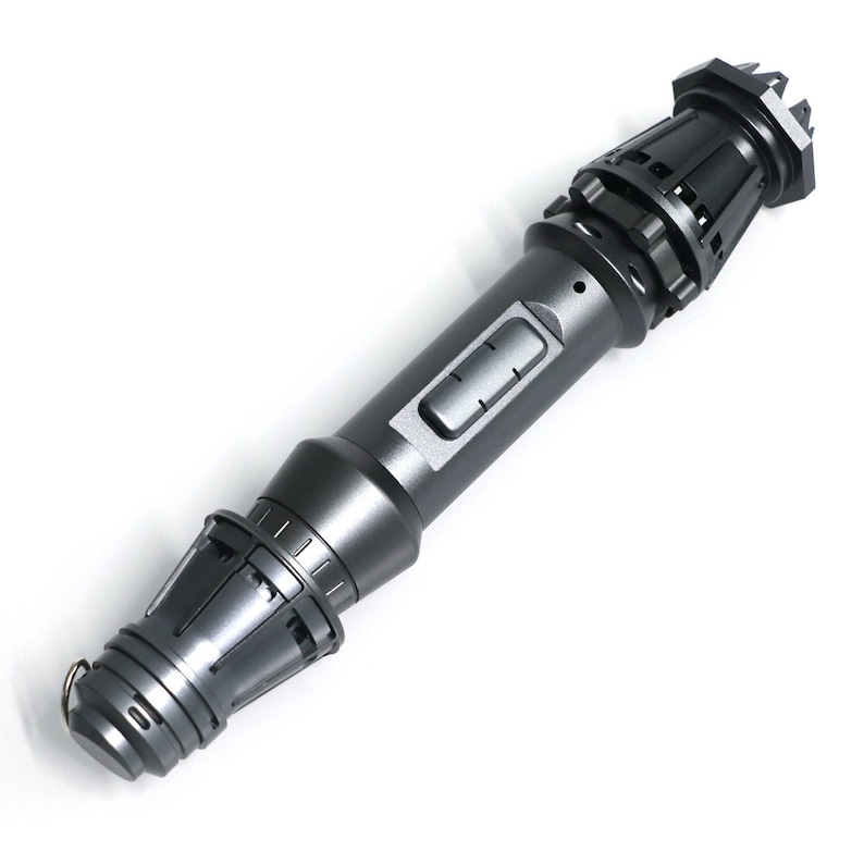 May include: A dark gray metal lightsaber with a textured surface and a black button. The lightsaber has a pointed tip and a rounded handle.
