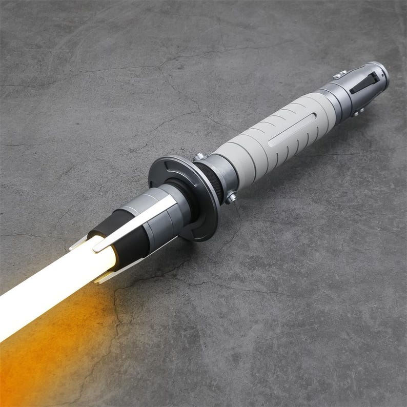 Peut inclure: Un sabre laser argent&eacute; avec une poign&eacute;e blanche et une lame orange vif. Le sabre laser est sur une surface grise.