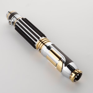 Puede incluir: Un sable de luz plateado y dorado con un agarre negro y plateado. El sable de luz tiene un agarre negro texturizado y un acento dorado alrededor del medio.