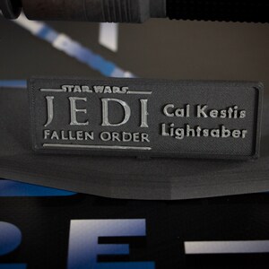May include: A black lightsaber replica with a silver plaque that reads "Star Wars Jedi Fallen Order Cal Kestis Lightsaber".