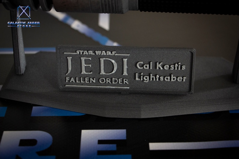May include: A black lightsaber replica with a silver plaque that reads "Star Wars Jedi Fallen Order Cal Kestis Lightsaber".