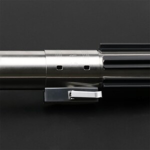 May include: A silver lightsaber hilt with a black handle and a silver switch. The hilt has a series of grooves and a small, circular opening on the side.