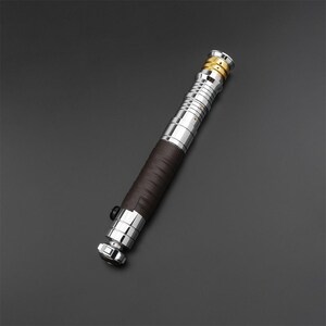 May include: A silver and brown lightsaber with a leather grip. The lightsaber has a gold accent at the top.