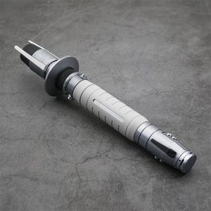 Peut inclure: Un sabre laser blanc et argent avec une poign&eacute;e noire et une lame argent&eacute;e. Le sabre laser est sur une surface grise.