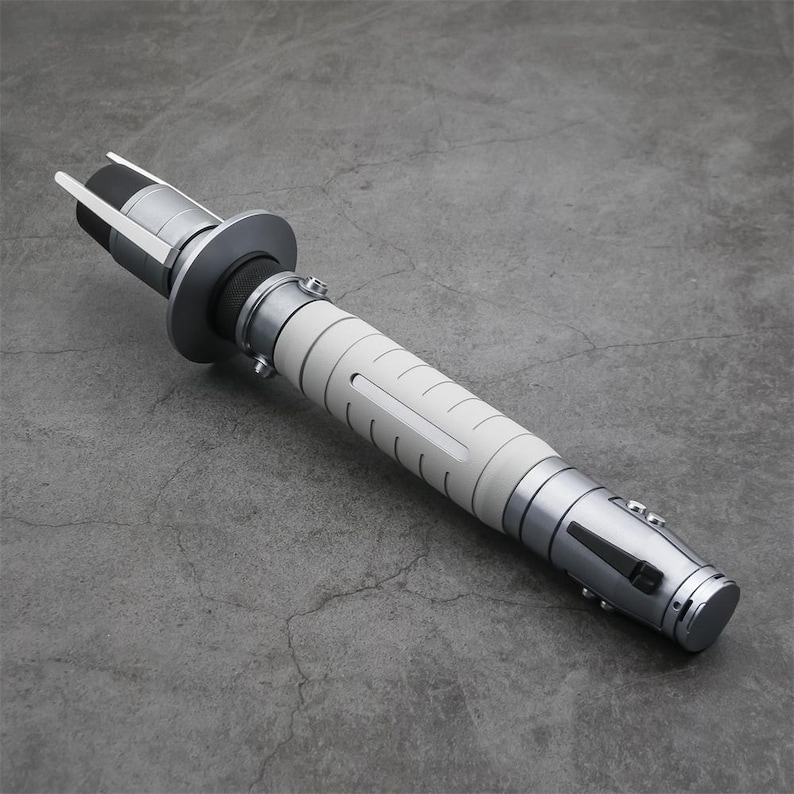 Peut inclure: Un sabre laser blanc et argent avec une poign&eacute;e noire et une lame argent&eacute;e. Le sabre laser est sur une surface grise.