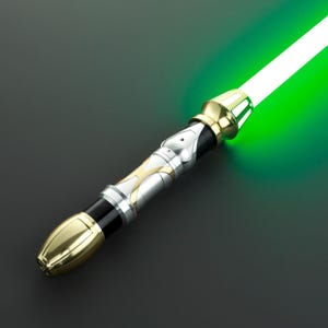 Puede incluir: Un sable de luz plateado y dorado con una hoja verde. El sable de luz tiene un diseño detallado con un mango negro.