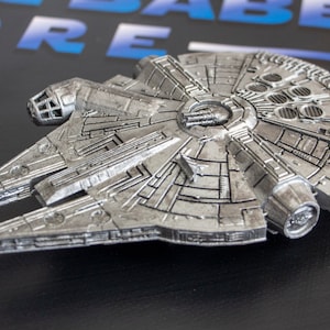 May include: A silver-colored model of the Millennium Falcon spaceship from Star Wars. The model is detailed with intricate lines and textures, and features a raised cockpit and engine exhaust ports.