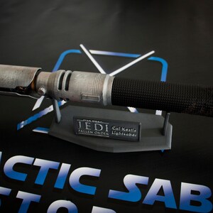 May include: A weathered, silver lightsaber with a black grip and a damaged emitter. The lightsaber is on a black stand with a silver plaque that reads "Jedi Cal Kestis Fallen Order Lightsaber".