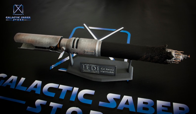 May include: A weathered, silver lightsaber with a black grip and a damaged emitter. The lightsaber is on a black stand with a silver plaque that reads "Jedi Cal Kestis Fallen Order Lightsaber".