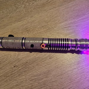 Peut inclure: Un sabre laser argent&eacute; avec des motifs grav&eacute;s complexes et une lame violette lumineuse. Le manche comporte des boutons noirs et un bouton orange &eacute;clair&eacute;. Le sabre laser est pos&eacute; sur une surface en bois.