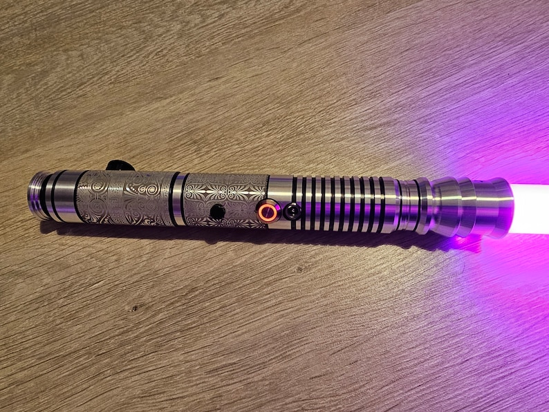 Peut inclure: Un sabre laser argent&eacute; avec des motifs grav&eacute;s complexes et une lame violette lumineuse. Le manche comporte des boutons noirs et un bouton orange &eacute;clair&eacute;. Le sabre laser est pos&eacute; sur une surface en bois.