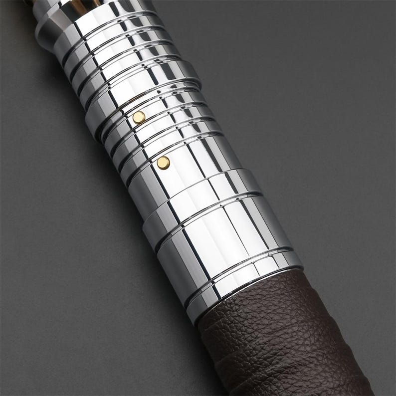 May include: Close-up of a silver lightsaber hilt with a brown leather grip. The hilt has a series of grooves and two small gold accents.