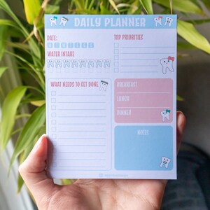 Puede incluir: Un planificador diario azul y rosa con un diseño de diente. El planificador tiene secciones para la fecha, las principales prioridades, lo que hay que hacer, el desayuno, el almuerzo, la cena y las notas.