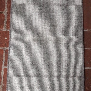 Puede incluir: Una alfombra rayada blanca y gris con flecos en todos los lados. La alfombra está hecha de un material tejido y tiene una textura sutil.