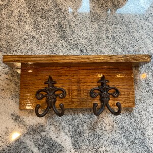 May include: A rustic wooden wall shelf with two ornate metal hooks. The shelf is made of light brown wood with a distressed finish. The hooks are dark brown and feature a decorative fleur-de-lis design.