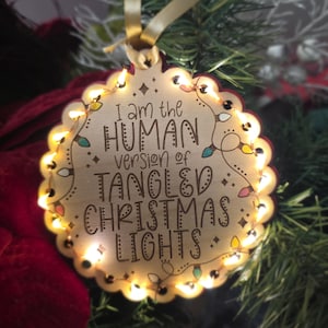 May include: Wooden Christmas ornament with the text "I am the HUMAN version of TANGLED CHRISTMAS LIGHTS." The ornament is round with a scalloped edge and is illuminated with small lights. A gold ribbon is attached for hanging.