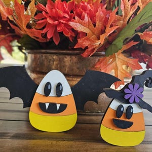 May include: Two wooden candy corn decorations, one with black bat wings and a vampire face, the other with a witch's hat and a smiling face.