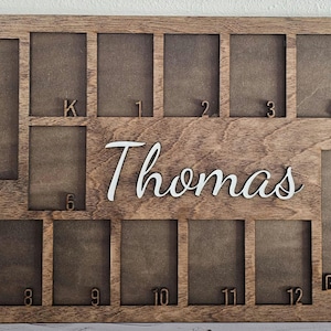 May include: A brown wooden photo frame with 13 slots for pictures. The slots are labeled with the grades Pre-K through 12 and Graduation. The frame has the name "Thomas" written in white lettering.