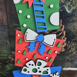 May include: A wooden Christmas decoration shaped like a stack of presents. The top present is green with white polka dots and a red bow. The middle present is red with green trees and a blue bow. The bottom present is blue with white snowflakes and a red and white striped scarf.