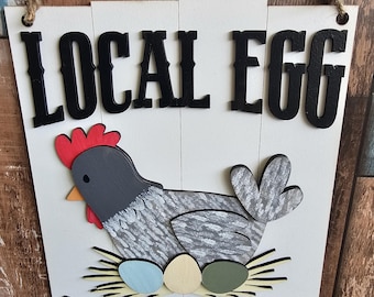 Puede incluir: Un letrero de madera blanco con letras negras que dice "LOCAL EGG DEALER". Una gallina gris y blanca con una cresta roja está sentada en un nido de paja con tres huevos de colores.