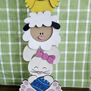 May include: A wooden Easter decoration with a yellow chick, a white lamb, and a white bunny holding a blue and white egg. The decoration is standing on a green base and there are five colorful striped Easter eggs on a wooden surface.