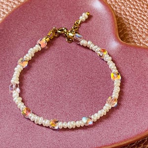 May include: A delicate bracelet featuring small, white pearls and iridescent, faceted crystal beads. The bracelet has a gold-colored clasp and is displayed on a pink heart-shaped dish, creating a feminine and elegant aesthetic.