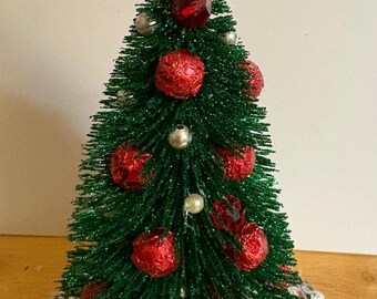 May include: A miniature Christmas tree with green, glittery needles, adorned with red ornaments and silver accents. The tree is topped with a red bow and a green wreath. The base has a striped pattern.