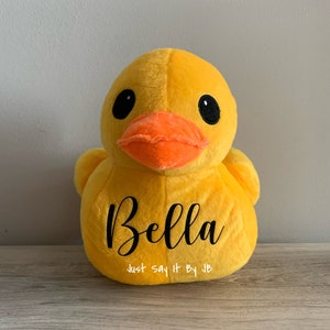May include: Yellow plush rubber duck toy with black embroidered eyes and an orange beak. The duck has the name "Bella" embroidered on its chest and the text "Just Say It By JB" underneath.