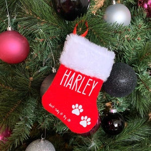May include: Red felt Christmas stocking with white faux fur trim. The stocking has the name "Harley" written in white lettering and the text "Just Say It By JB" with two white paw prints.