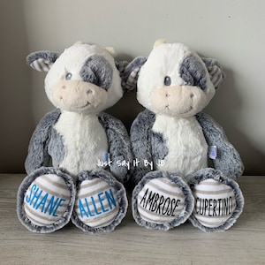 May include: Two plush gray and white cow stuffed animals with blue and gray striped feet. The feet are personalized with the names "Shane Allen" and "Ambrose Cupertino".