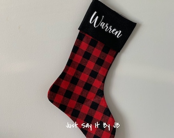 May include: A Christmas stocking with a red and black buffalo plaid pattern. The top of the stocking is black with the name "Warren" in white script. A black loop is attached for hanging. The text "Just say it By JB" is at the bottom.