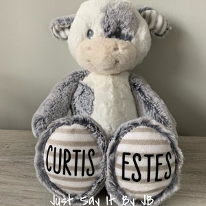 May include: A plush gray and white cow with black eyes and a pink nose. The cow is sitting with its legs crossed and has the names "Curtis Estes" written on its feet. The text "Just Say It By JB" is written below the feet.
