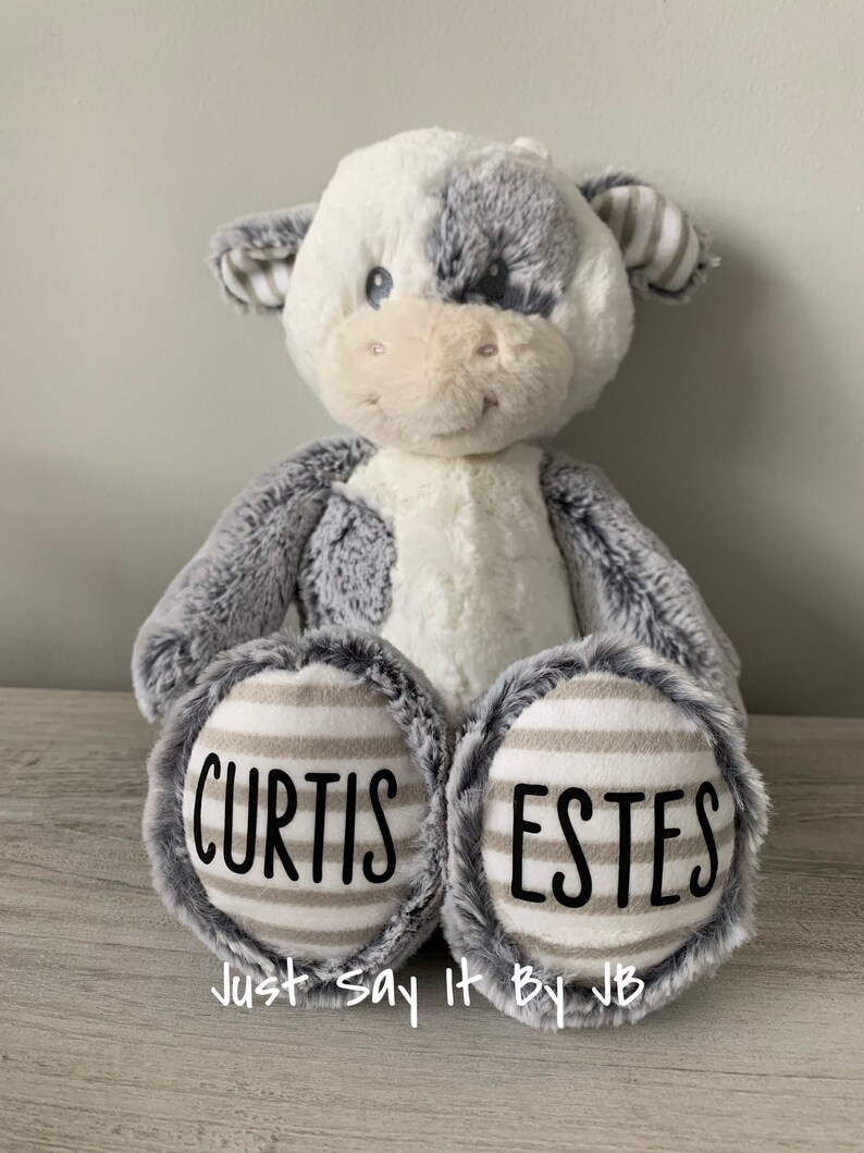May include: A plush gray and white cow with black eyes and a pink nose. The cow is sitting with its legs crossed and has the names "Curtis Estes" written on its feet. The text "Just Say It By JB" is written below the feet.