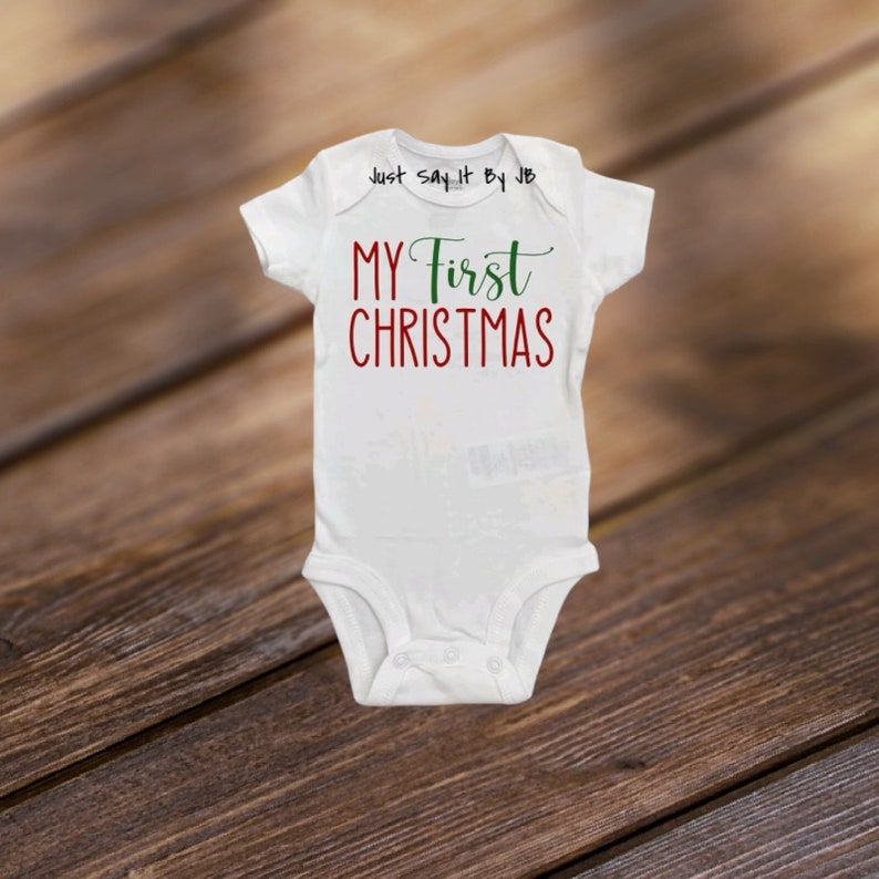 May include: White baby onesie with the text "MY First CHRISTMAS" in red and green. The onesie has short sleeves and a snap closure. The text "Just Say It By JB" is printed above the text.