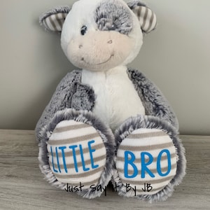 May include: A plush gray and white cow with a striped brown and white pattern on its ears. The cow has a blue embroidered smile and the words "LITTLE BRO" on its feet.