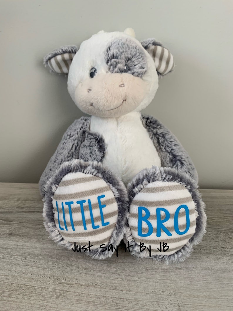 May include: A plush gray and white cow with a striped brown and white pattern on its ears. The cow has a blue embroidered smile and the words "LITTLE BRO" on its feet.