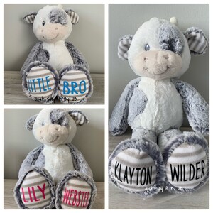 May include: Three plush cow toys with personalized names on the bottom of their feet. The first cow has the names "Little Bro" on its feet. The second cow has the names "Klayton Wilder" on its feet. The third cow has the names "Lily Webster" on its feet. All three cows have gray and white fur.