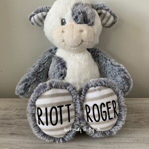 May include: A plush gray and white cow stuffed animal with black and white striped ears. The cow has a gray patch over one eye. The cow is sitting with its legs crossed and has the names "RIOTT" and "ROGER" embroidered on its feet.