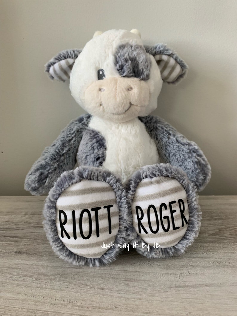 May include: A plush gray and white cow stuffed animal with black and white striped ears. The cow has a gray patch over one eye. The cow is sitting with its legs crossed and has the names "RIOTT" and "ROGER" embroidered on its feet.