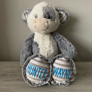 May include: A plush cow toy with gray and white fur. The cow has a blue embroidered smile and eyes. The bottom of the cow's feet are gray and white striped fabric with the names "Hunter Wayne" embroidered in blue. The text "Just Say It By JB" is also embroidered in blue on the bottom of the cow's feet.