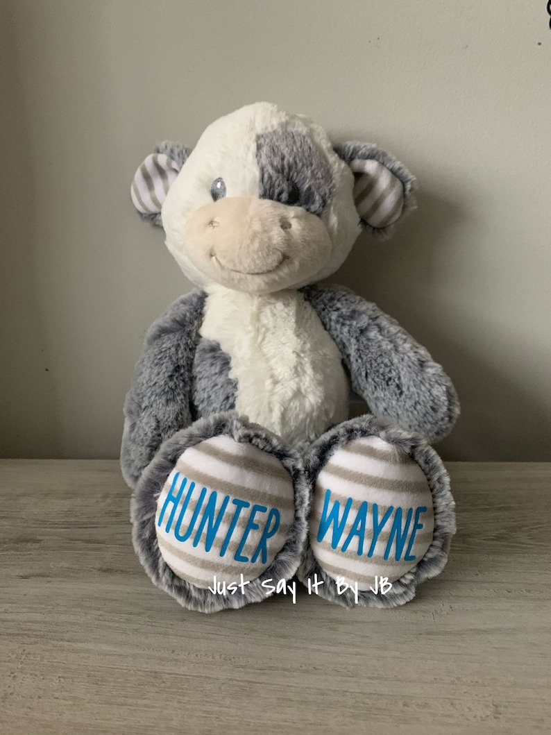 May include: A plush cow toy with gray and white fur. The cow has a blue embroidered smile and eyes. The bottom of the cow's feet are gray and white striped fabric with the names "Hunter Wayne" embroidered in blue. The text "Just Say It By JB" is also embroidered in blue on the bottom of the cow's feet.