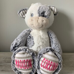 May include: A white and gray plush cow with black eyes and a pink nose. The cow has a gray and white striped pattern on its ears and feet. The words "MADISON" and "CORRINA" are embroidered on the feet in pink.