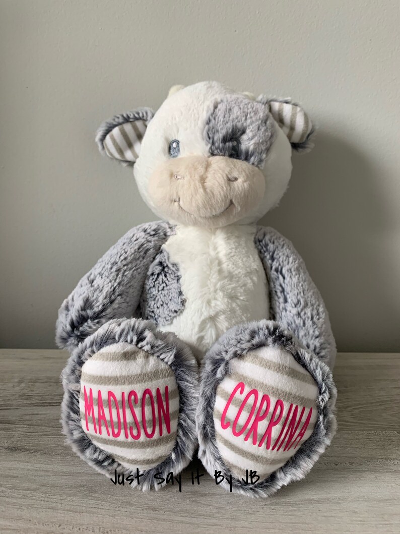 May include: A white and gray plush cow with black eyes and a pink nose. The cow has a gray and white striped pattern on its ears and feet. The words "MADISON" and "CORRINA" are embroidered on the feet in pink.