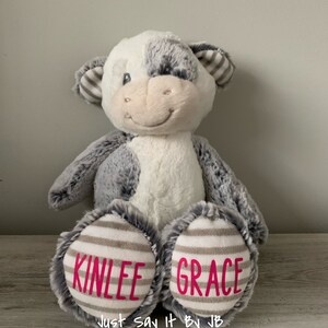 May include: A plush gray and white cow stuffed animal with the names "Kinlee" and "Grace" embroidered on its feet. The text "Just Say It By JB" is printed below the names.