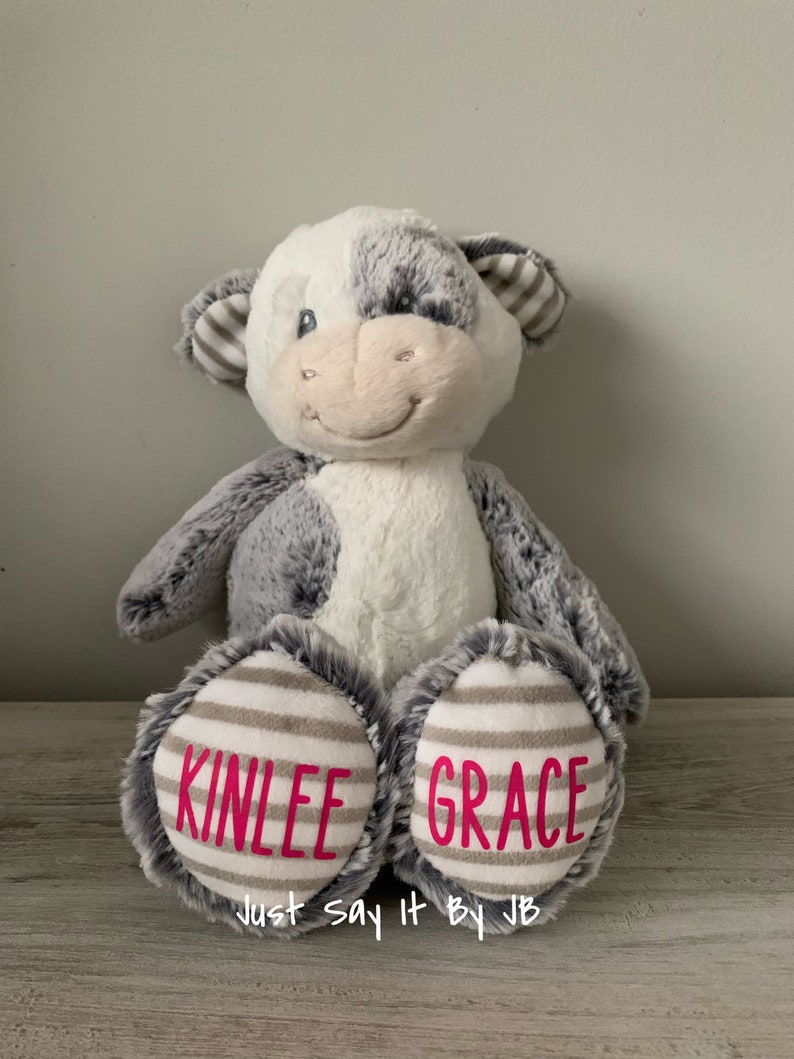 May include: A plush gray and white cow stuffed animal with the names "Kinlee" and "Grace" embroidered on its feet. The text "Just Say It By JB" is printed below the names.