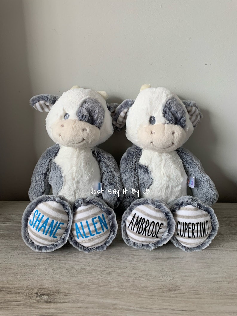 May include: Two plush gray and white cow stuffed animals with blue and gray striped feet. The feet are personalized with the names "Shane Allen" and "Ambrose Cupertino".