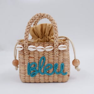 Könnte beinhalten: Eine kleine Strohtasche mit einem blauen "Bleu" bestickt auf der Vorderseite. Die Tasche hat einen weißen Muschelbesatz und Holzperlenakzente.