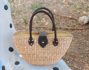 May include: A woven straw tote bag with brown leather handles and a brown leather flap closure. The bag has a zipper closure and a floral design on the flap.