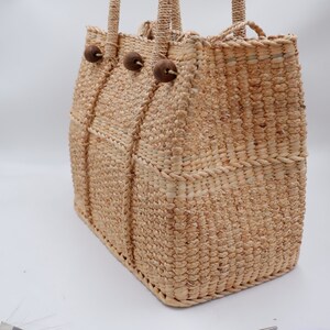 K&ouml;nnte beinhalten: Eine geflochtene Strohtasche mit Holz-Knopf-Akzenten. Die Tasche hat eine nat&uuml;rliche beige Farbe und eine rechteckige Form.