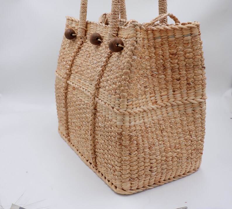 K&ouml;nnte beinhalten: Eine geflochtene Strohtasche mit Holz-Knopf-Akzenten. Die Tasche hat eine nat&uuml;rliche beige Farbe und eine rechteckige Form.